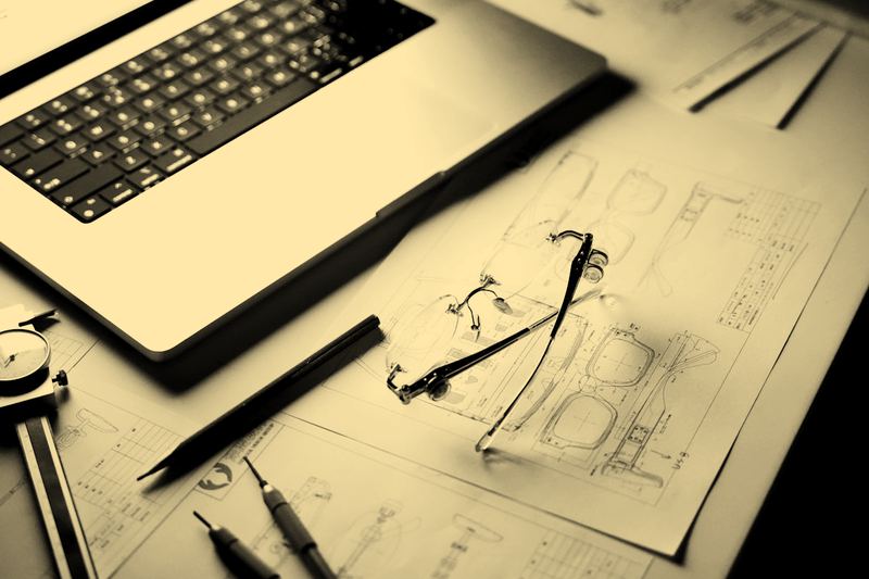 A desk at night with a laptop, technical drawings, drafting compasses and a pair of glasses, no person in frame