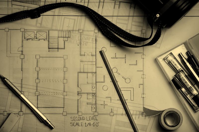 An architectural floor plan on a desk with drafting pencils, a metal ruler and a rolled drawing tube, no people in frame