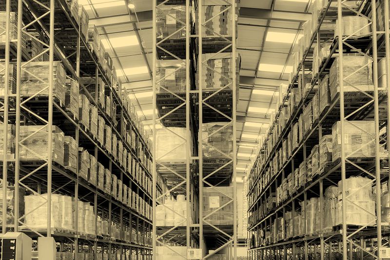 A wide aisle in a distribution warehouse between tall red and grey racking loaded with pallets, no people